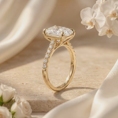 Side-profile view of a gold-tone cushion rectangular pavé solitaire ring beside white flowers.