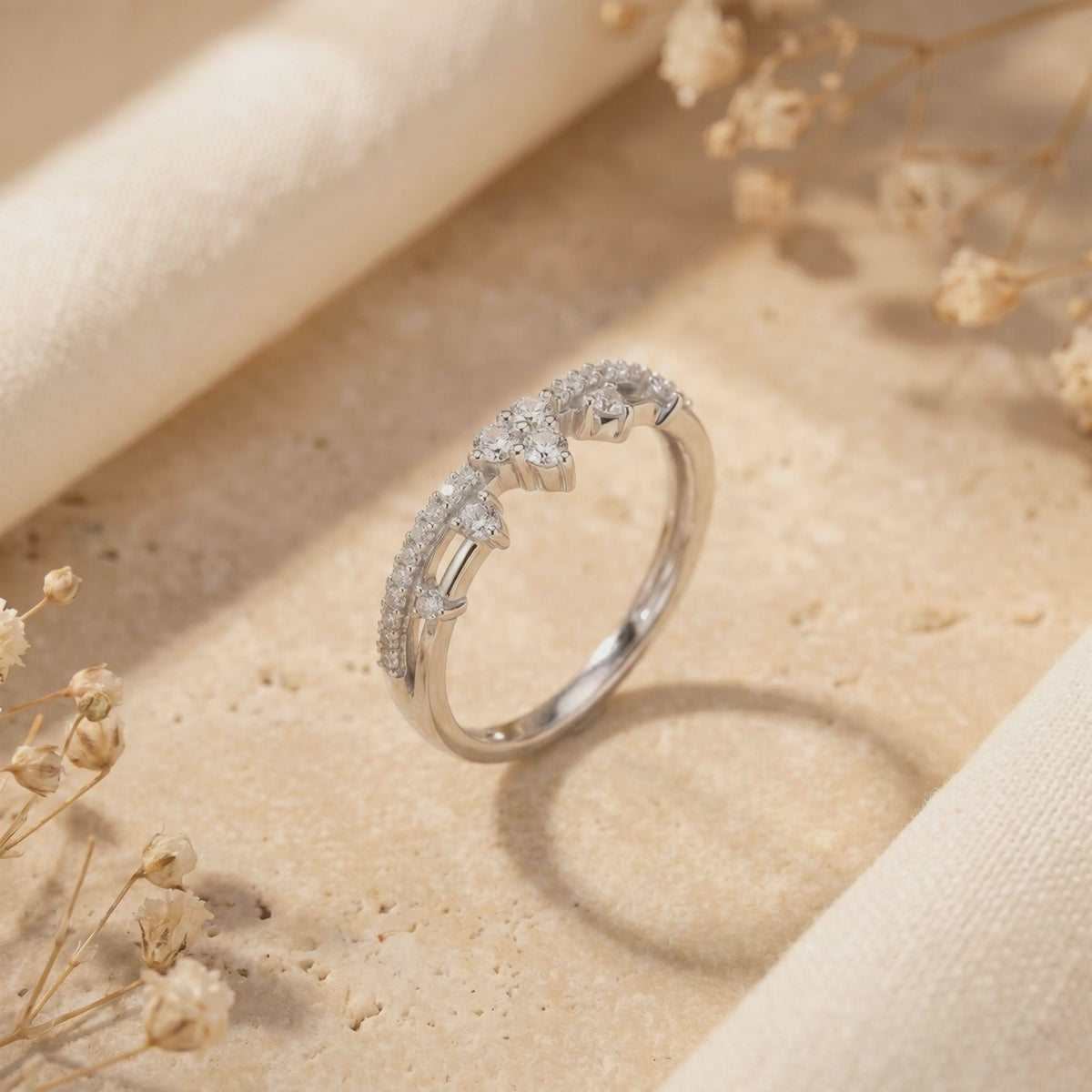 Tiara-style ring with clear stones displayed upright on a textured stone surface beside beige fabric and dried flowers.